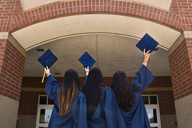 three women holding up graduation caps in front of college building