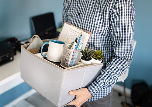 possible caucasian male in plaid shirt, holding box of office supplies and decorations, leaving his office after losing job