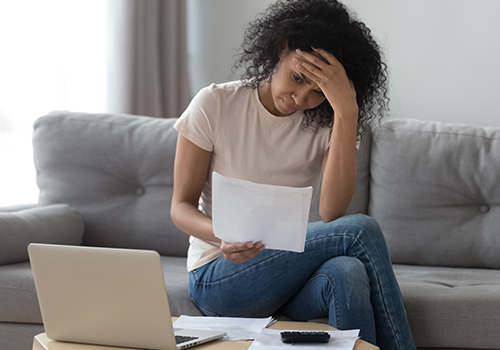 light skinned woman on couch worriedly reading paperwork in front of open laptop